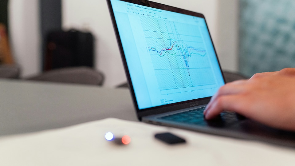 Hands typing on a laptop displaying a multicolored line graph on its screen, with a blurred USB drive or small device on the table in the foreground.